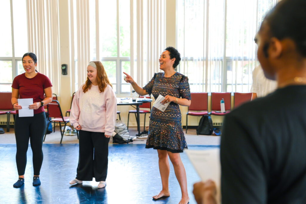 Four people stand inside a well-lit room. One person smiles while pointing her finger to instruct the class.