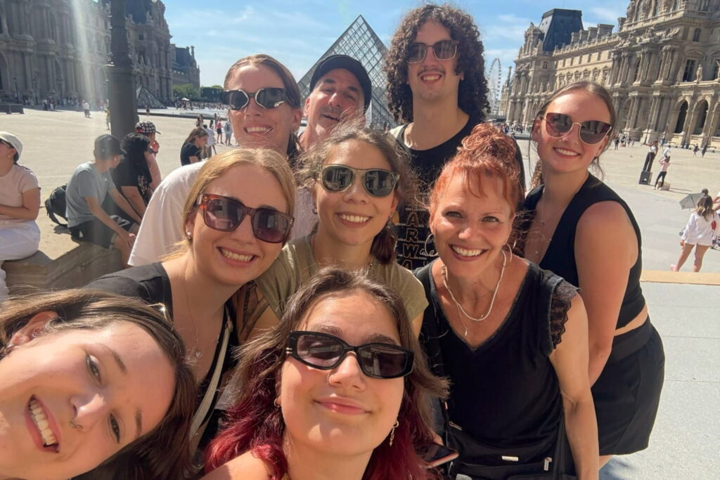 Nine people smile at the camera while standing outside of the Louvre Museum's Glass Pyramid in Paris, France.