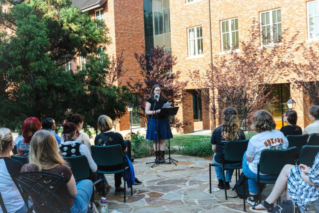 One person reads a paper to a class of peers outside in front of a brick-walled courtyard.
