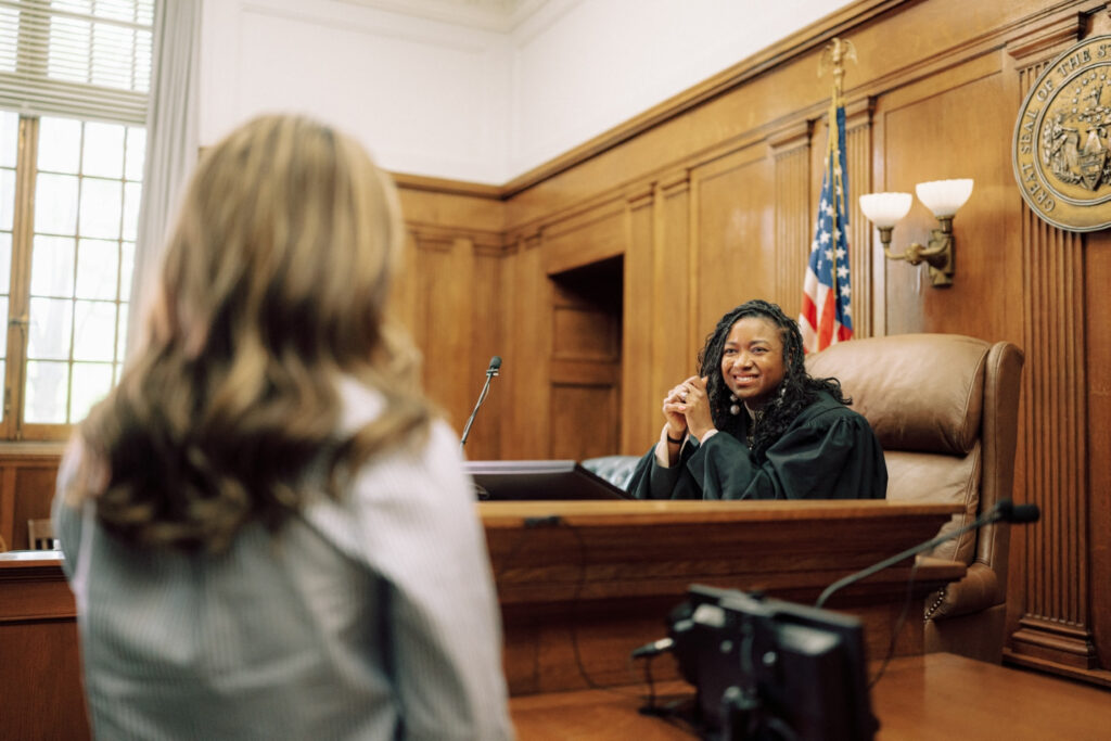 One person in a black gown sits at a judges bench with the United States flag behind them. The other person is standing facing the judge in conversation.