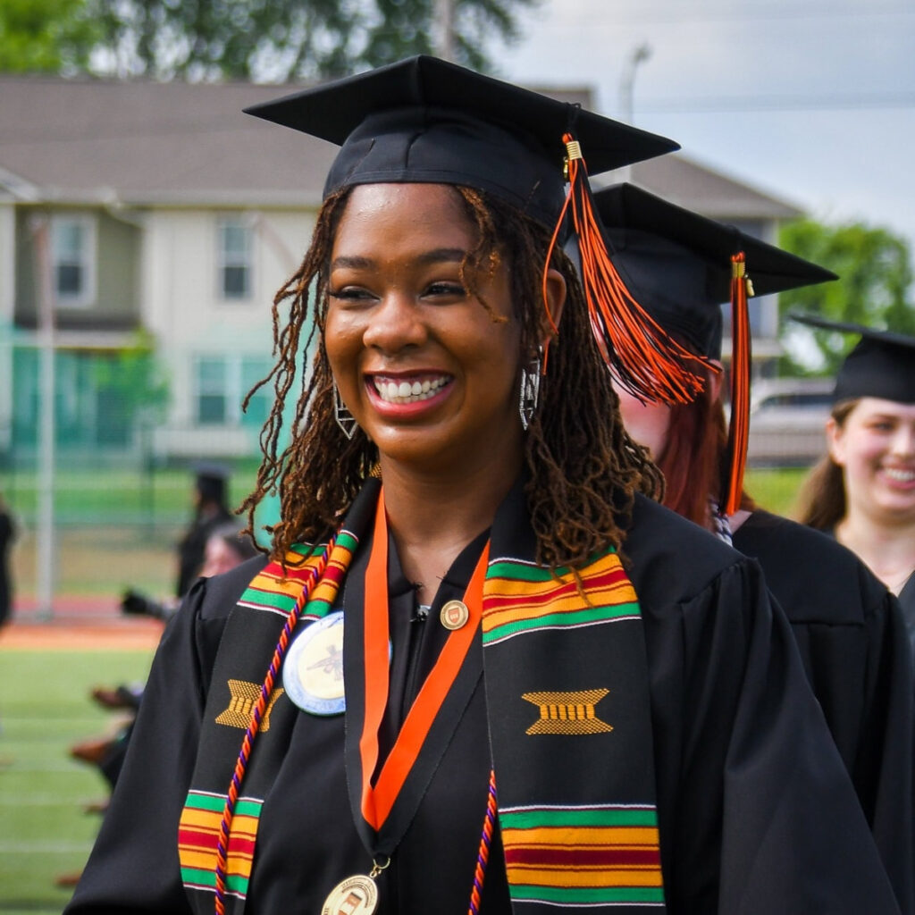 A smiling Hendrix College graduate is dressed in her cap, gown, and mortar board preparing to walk the stage at commencement.