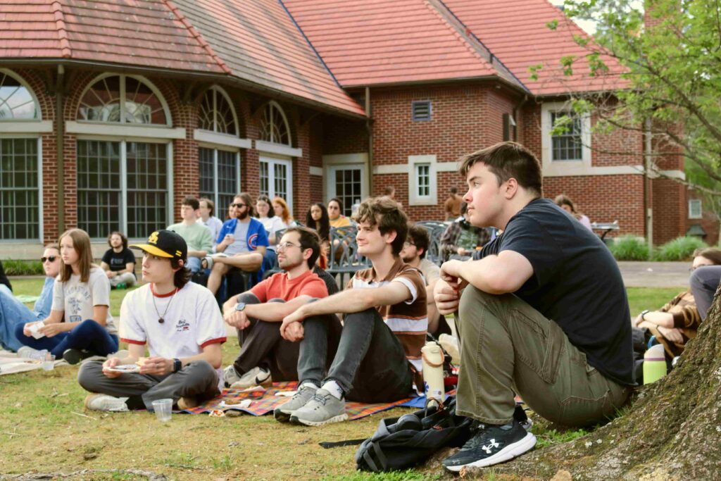 A crowd of more than a dozen students sit on the grass and on blankets at the back patio of the Bertie Wilson Murphy building, looking into the distance at a speaker who is not shown.