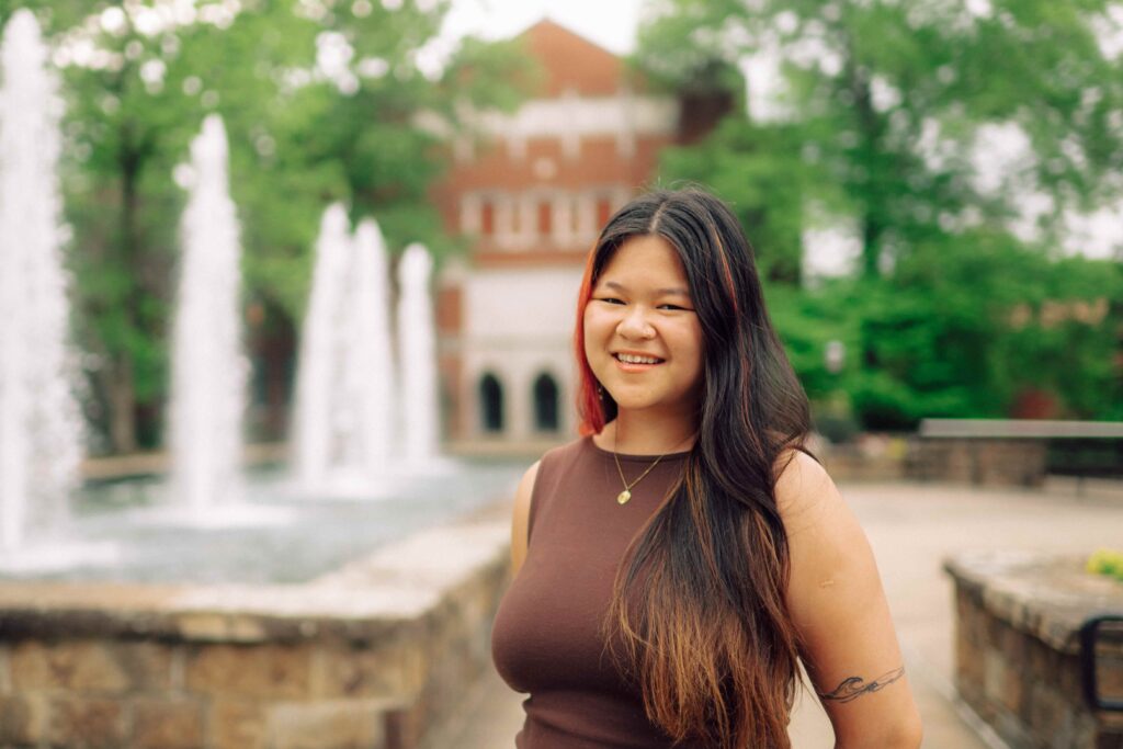A female student smiles while standing in front of the Hendrix fountain and the Staples Auditorium building on a sunny day.