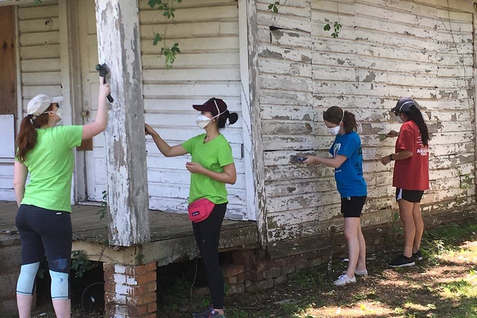 Four volunteers paint an old white house while wearing masks.