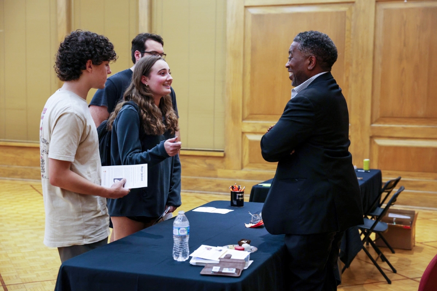Three students talk with an author at a book-signing event. A female student who is speaking gestures to the author who listens while smiling.