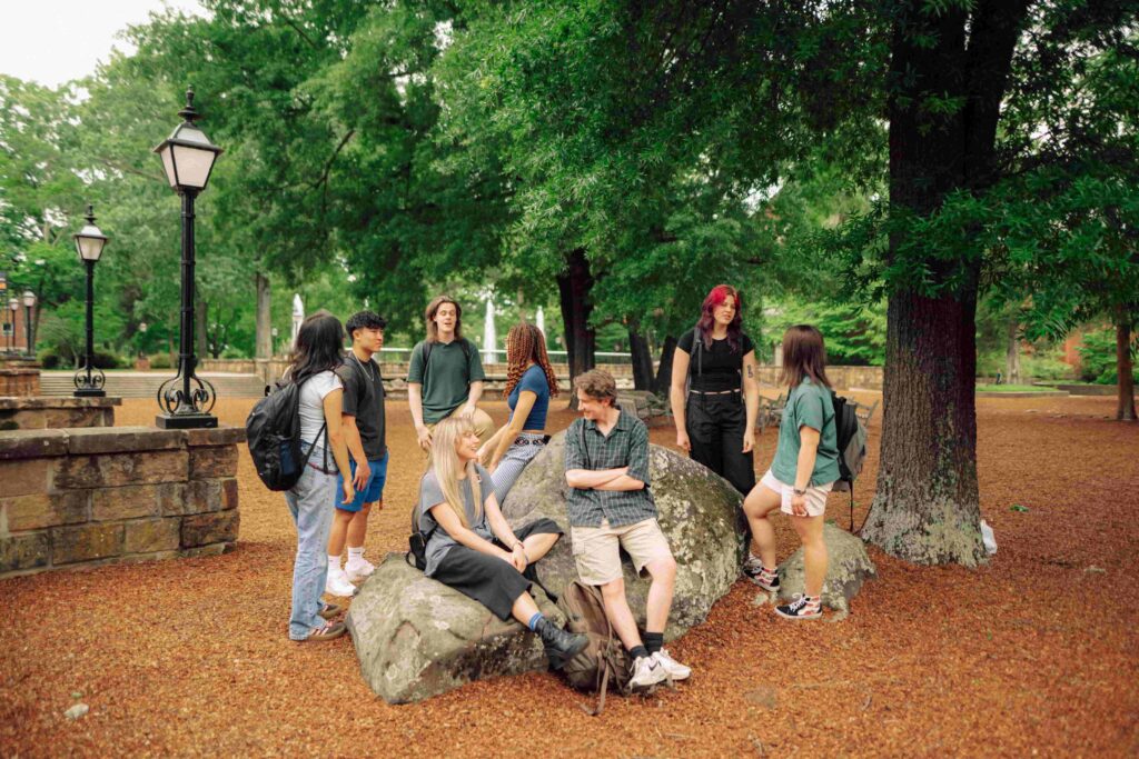 Eight students talk to each other beneath the trees of the Pecan Court at Hendrix College. Some stand while others sit on a large boulder.