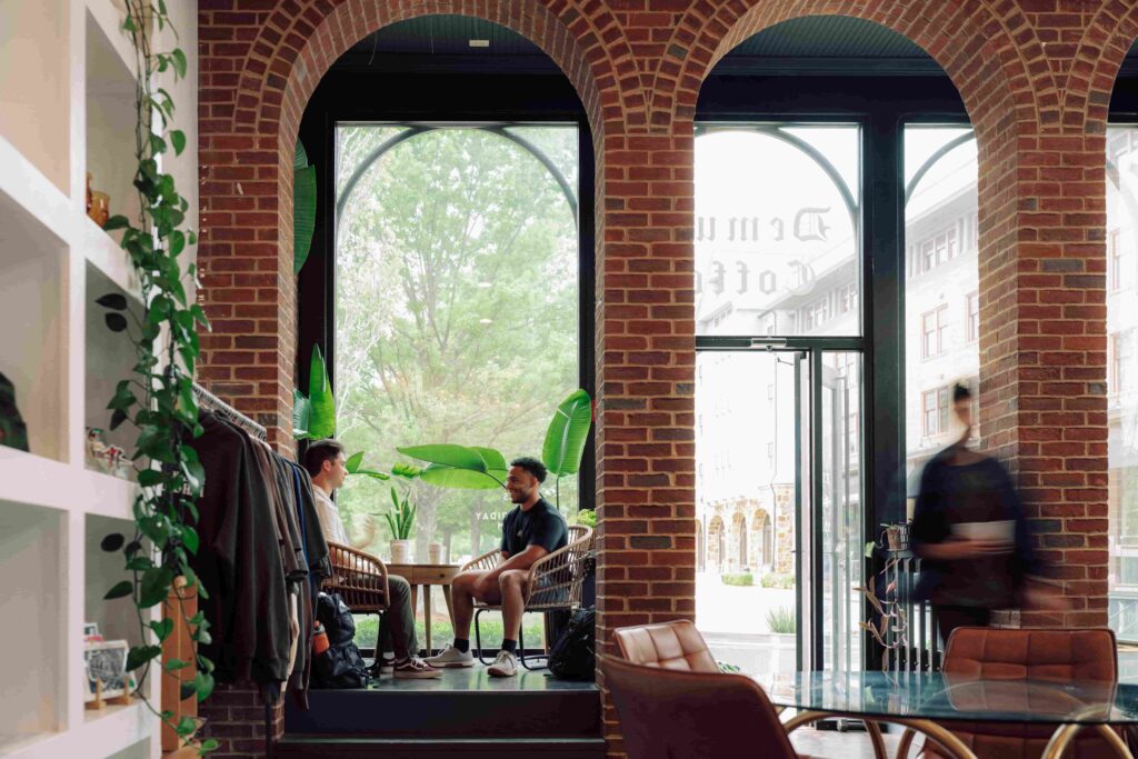 Students sitting in and moving through the lobby of a building with brick columns and large glass windows.
