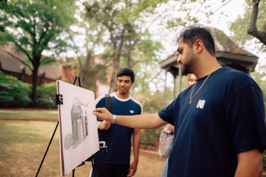 A student working on still life art outdoors with two other students watching.
