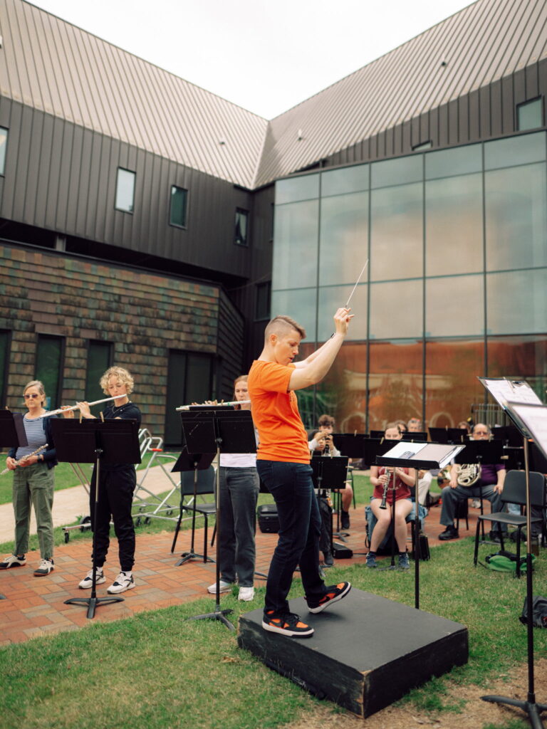A conductor leading a concert band rehearsing outdoors.