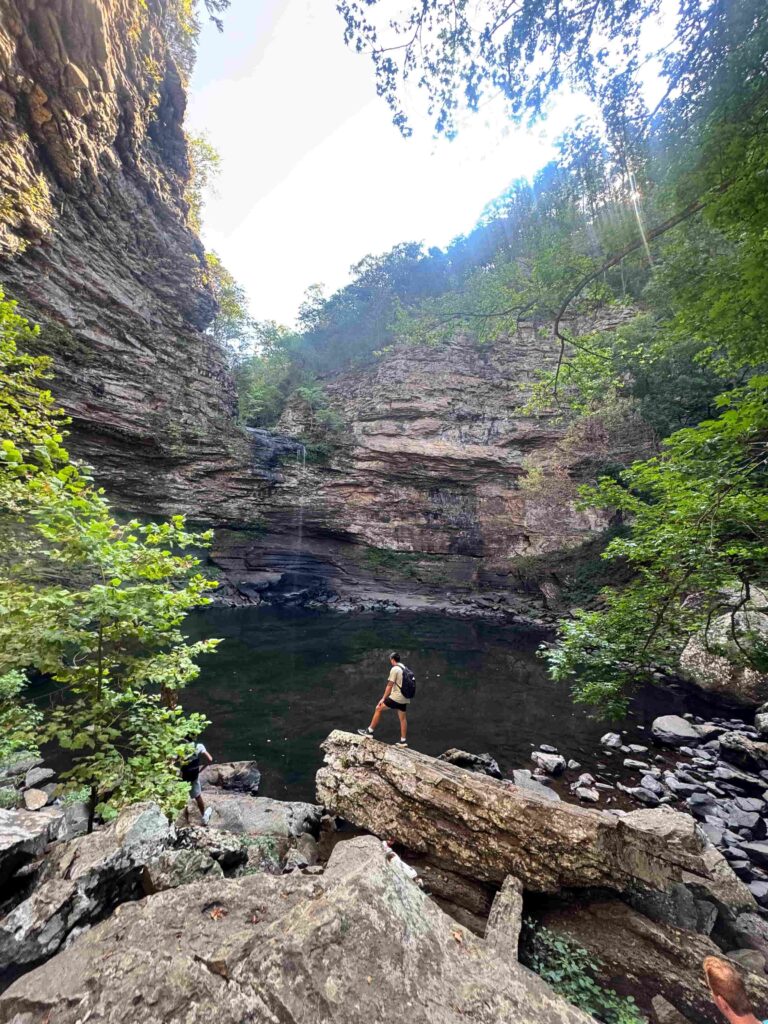 A student standing on a rock outcropping in a small ravine with a waterfall and lake.