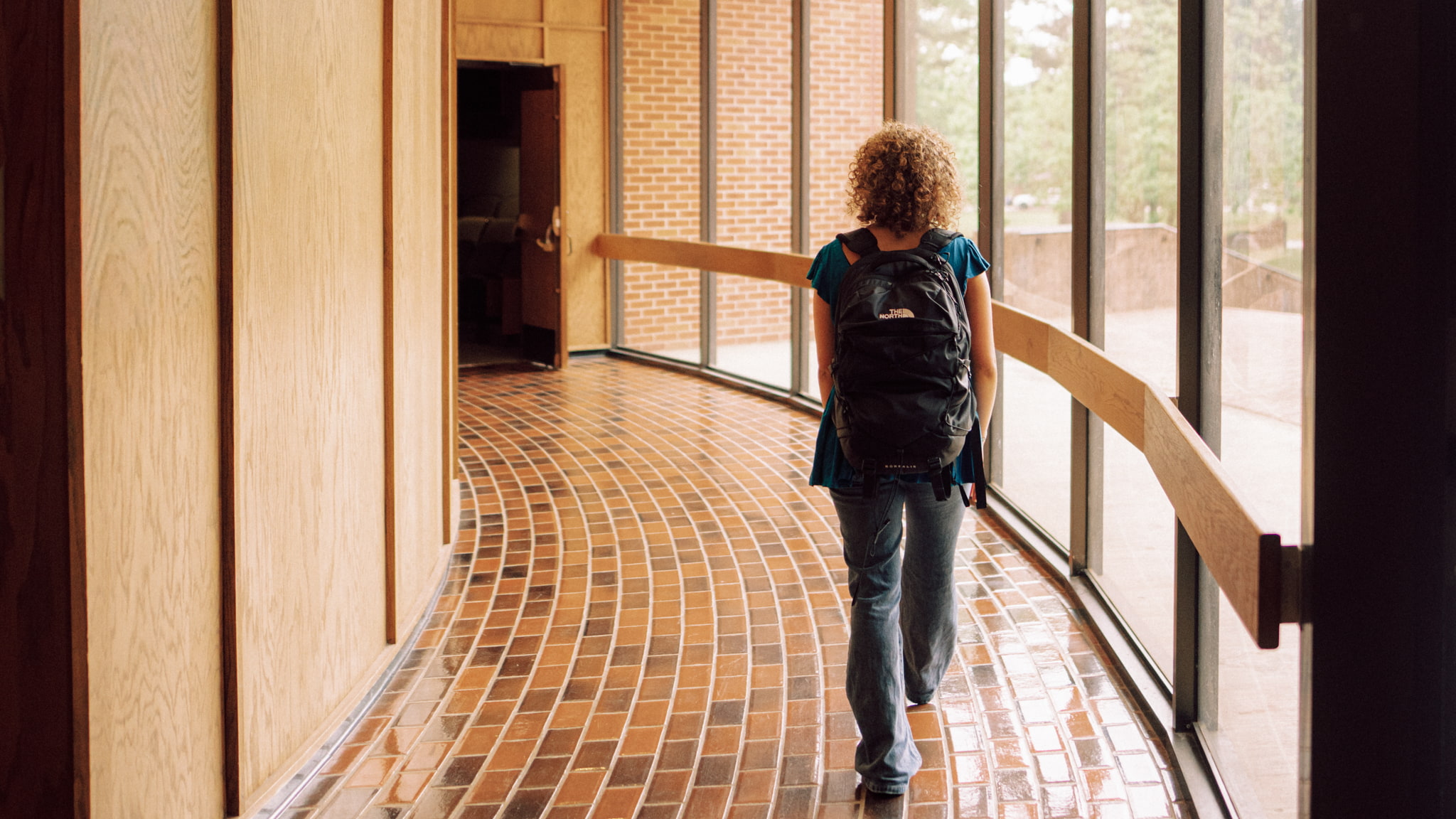 A student wearing a backpack walks down a curved glass-walled hallway with tile flooring on the Hendrix College campus.