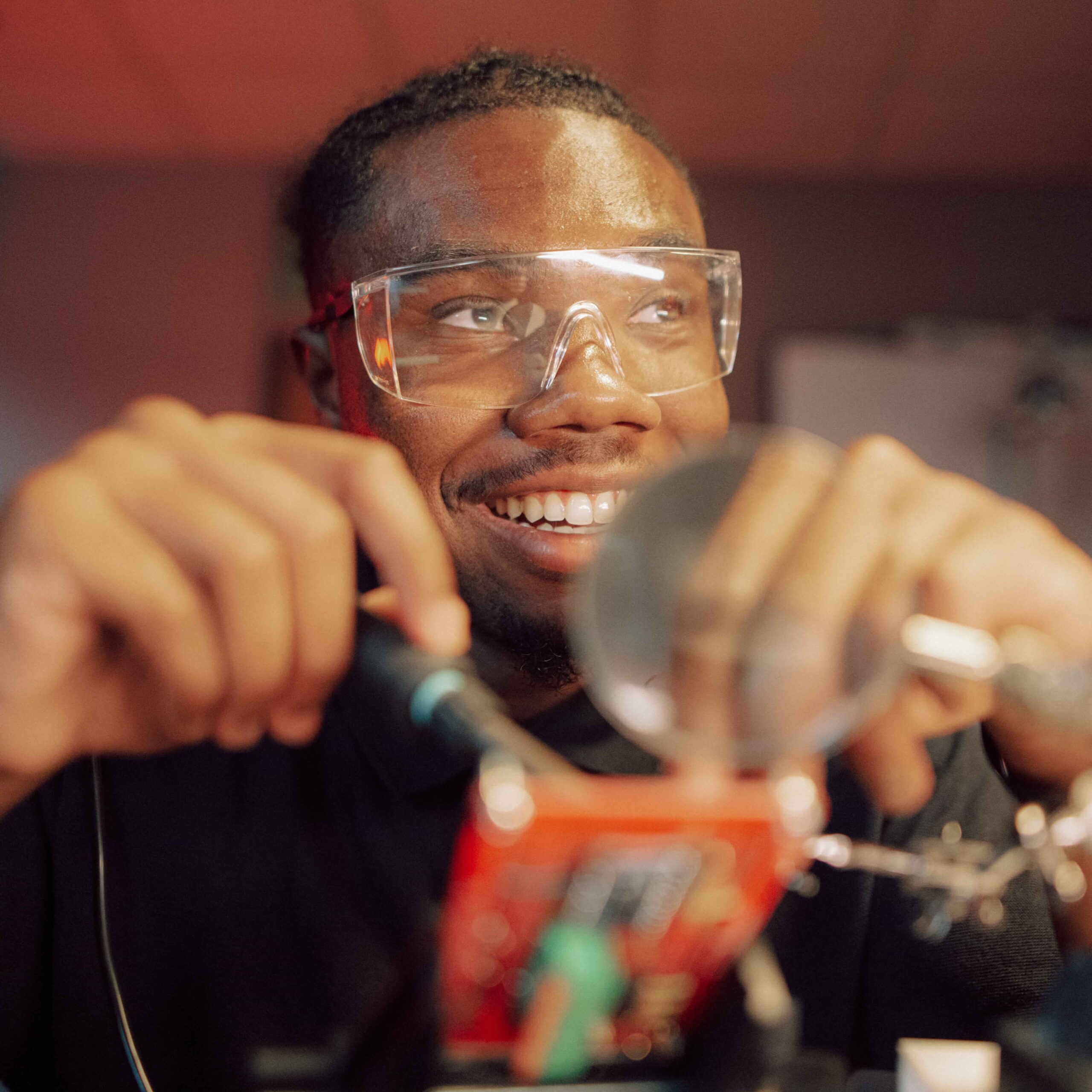 A smiling student in safety goggles looks up from working on a project.