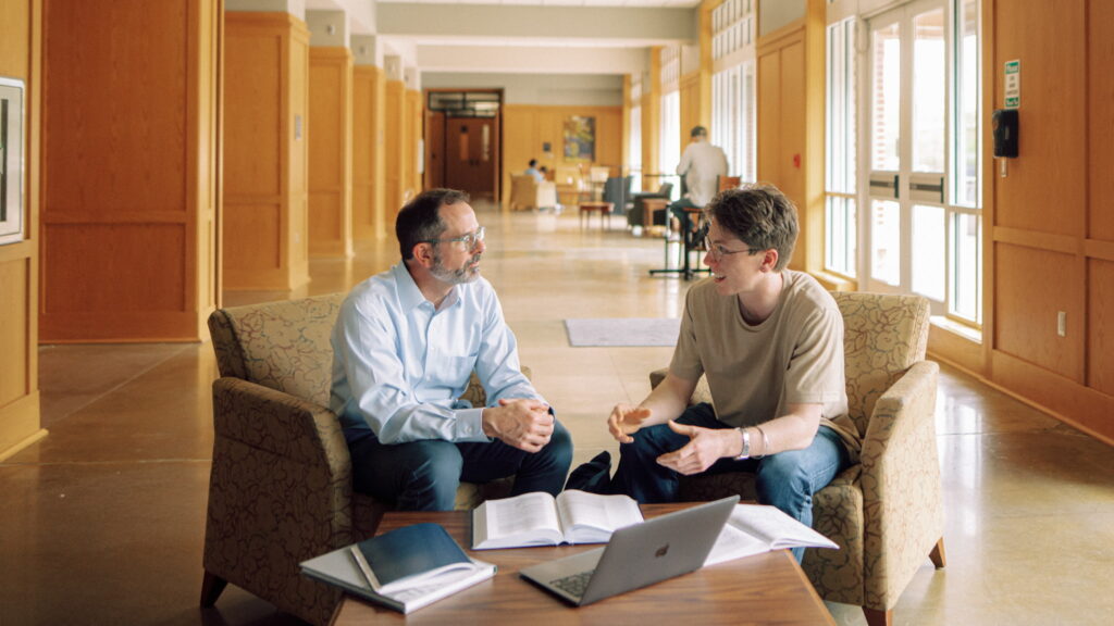 An academic advisor and Hendrix College student talk in comfortable chairs with books and a laptop in a campus hallway.