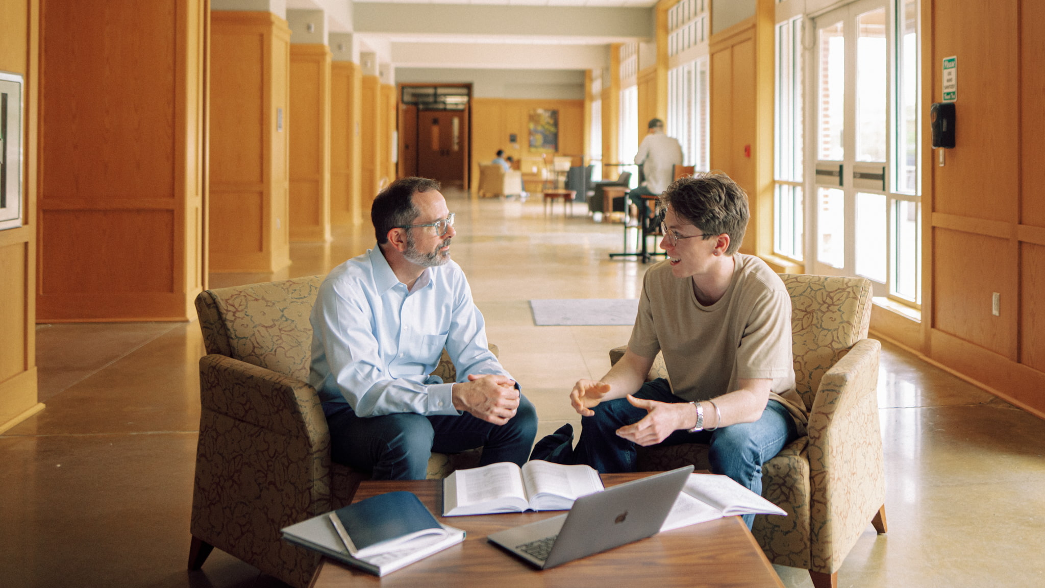 An academic advisor and Hendrix College student talk in comfortable chairs with books and a laptop in a campus hallway.