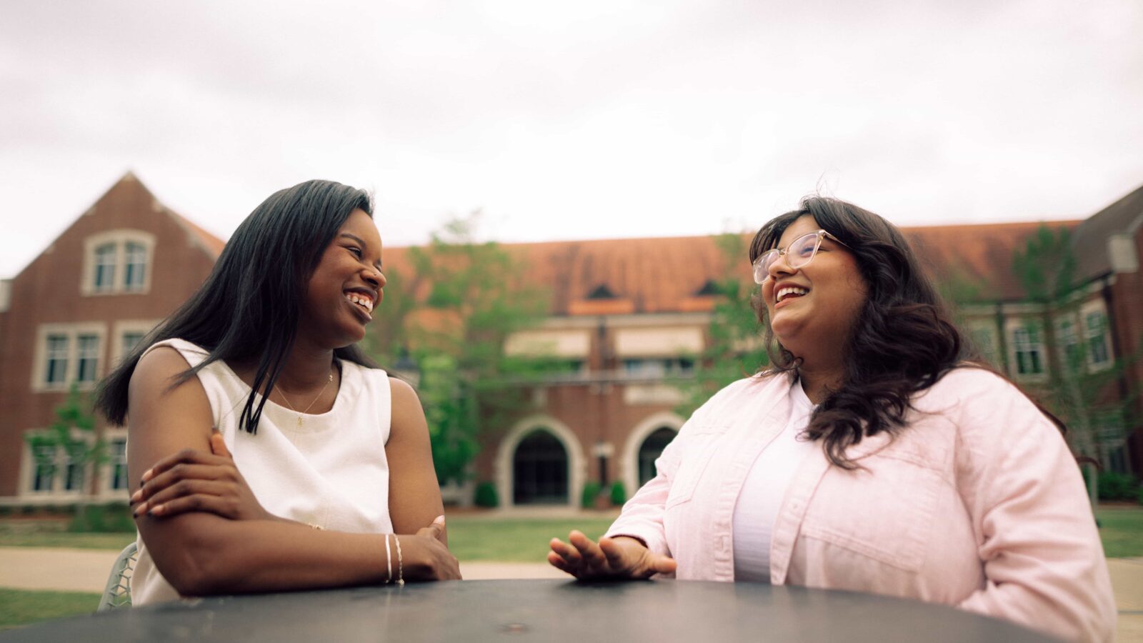 Two smiling Hendrix College students sit at an outdoor table with a large brick campus building in the background.