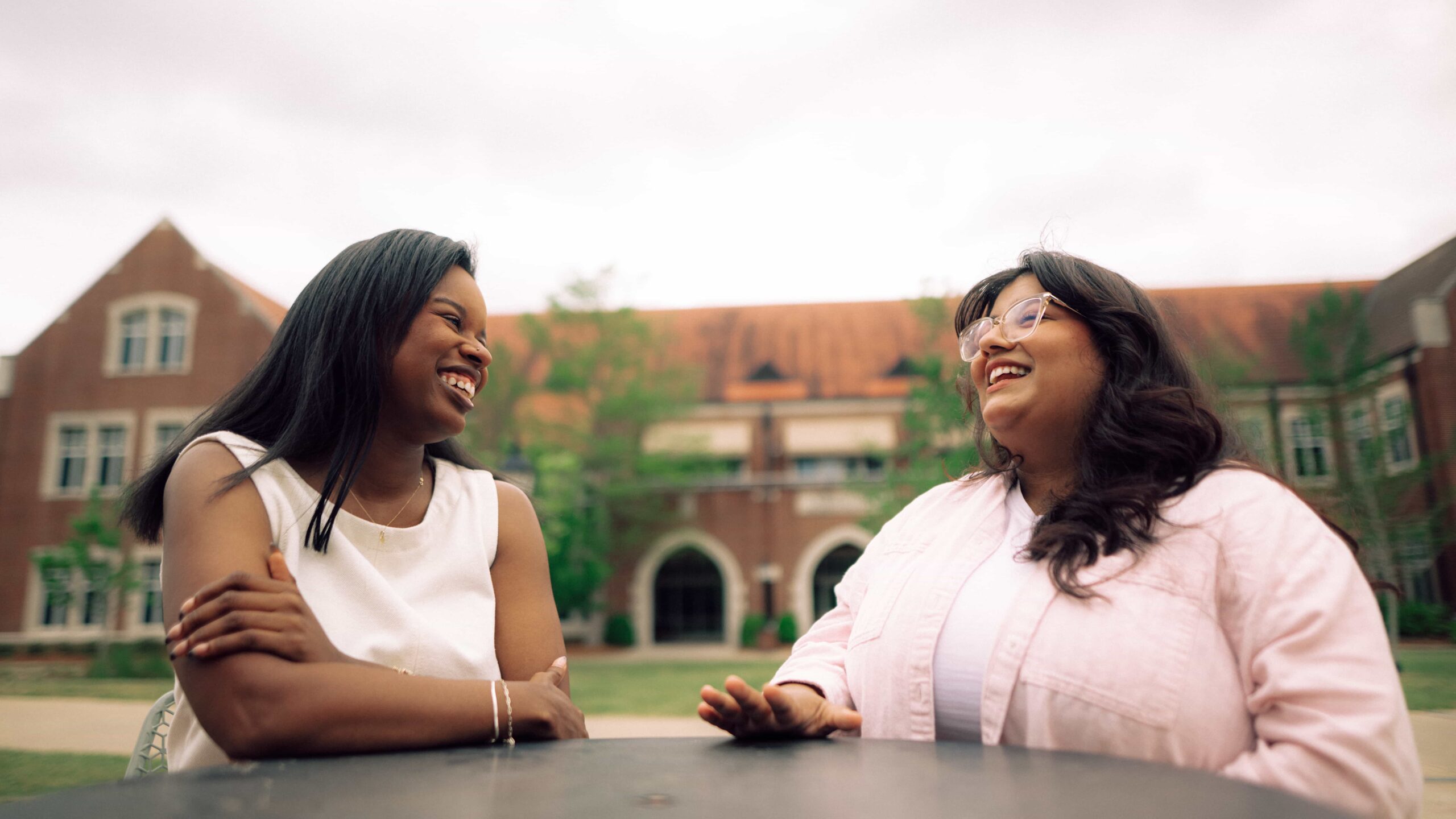 Two smiling Hendrix College students sit at an outdoor table with a large brick campus building in the background.