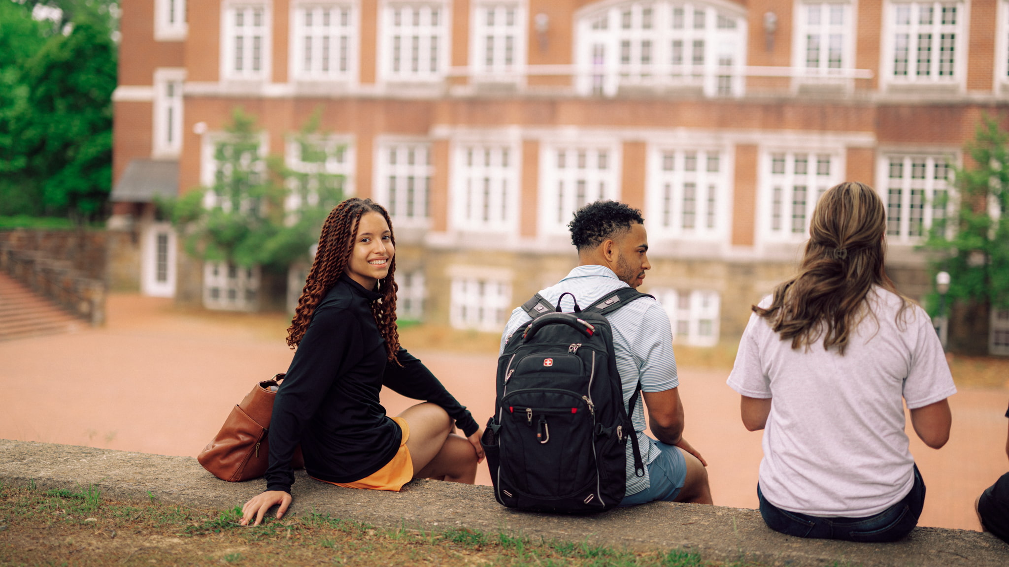 Three prospective Hendrix College students with backpacks sit outdoors on a low wall in front of a brick campus building.