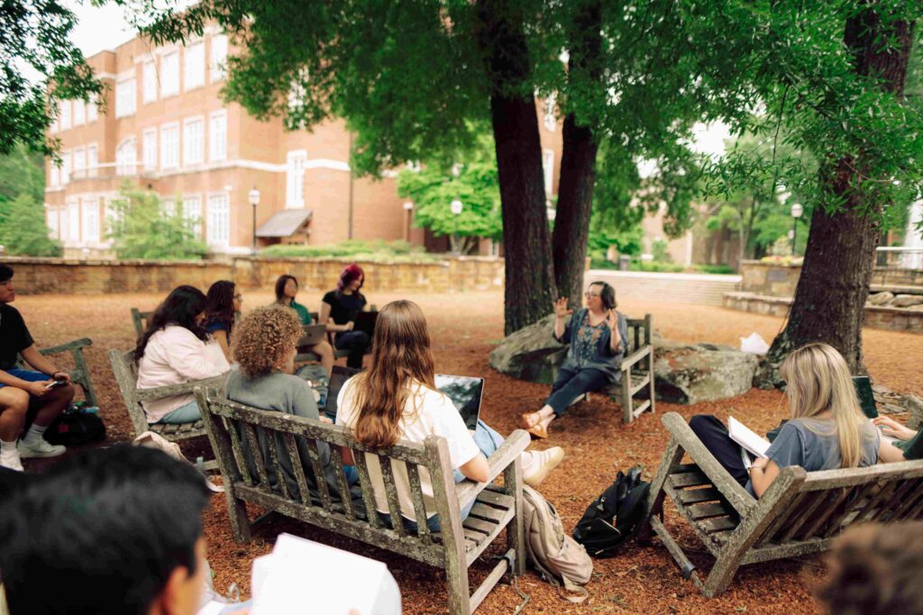 Students sitting on wooden benches in an outdoor classroom listening to a lesson from a faculty member.