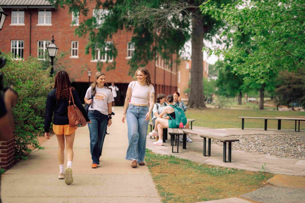 Students walking on a sidewalk on campus, with other students sitting on nearby benches.