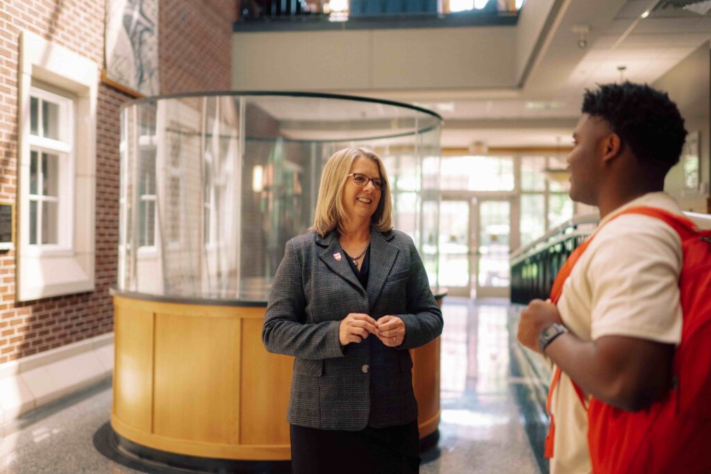 President Petersen speaking with a student in the hallway of a building on campus.