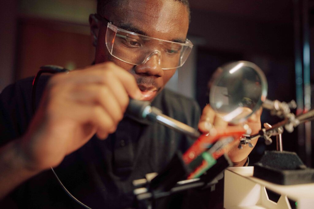 A male student wearing safety goggles soldering electronics components.