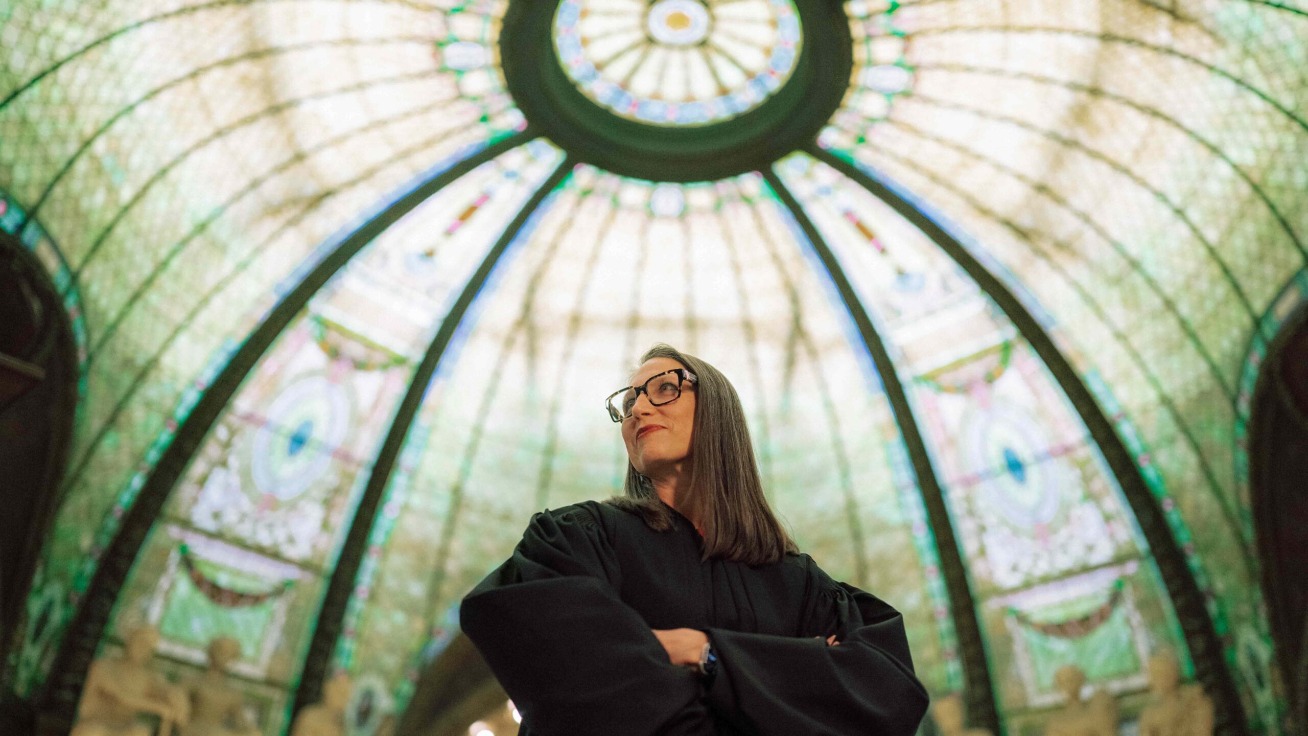 A Hendrix College alumni wearing black judicial robes stands looking up beneath a large ornate stained glass dome ceiling.