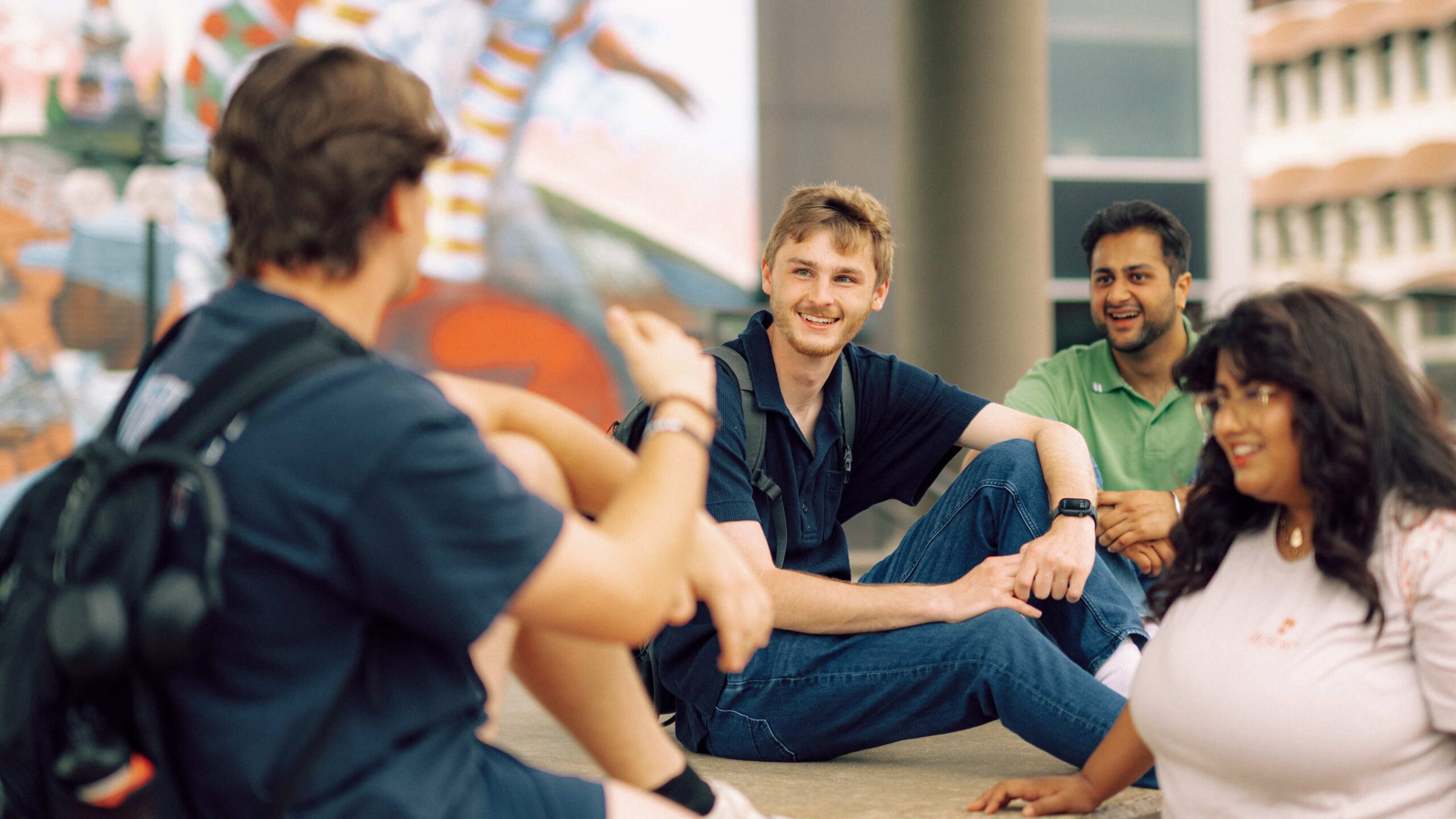 A group of Hendrix College students smile and chat together outdoors near an urban mural landscape in Conway, Arkansas.