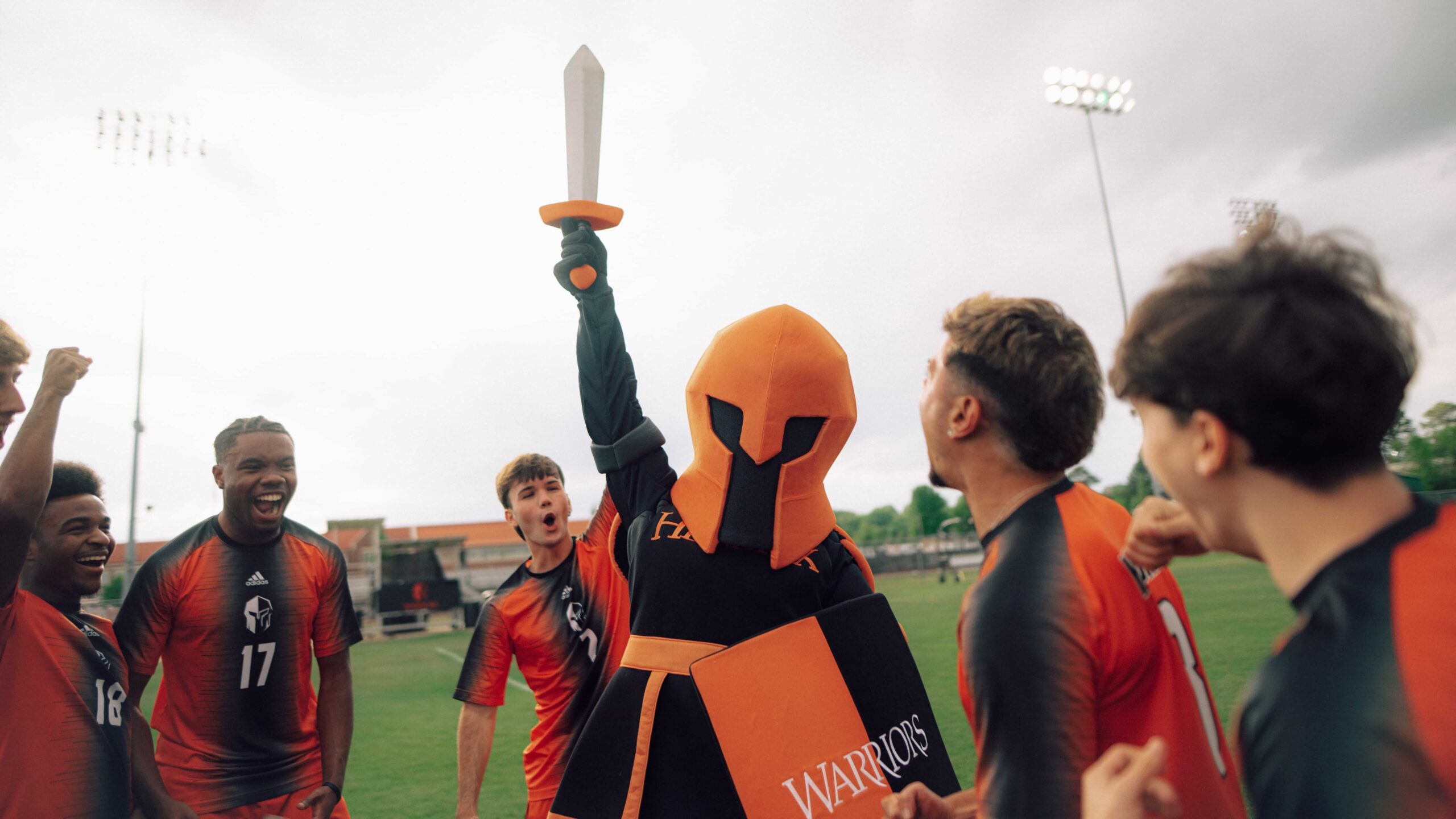 The Hendrix College Warriors mascot holds up a sword while celebrating on a sports field with soccer athletes.