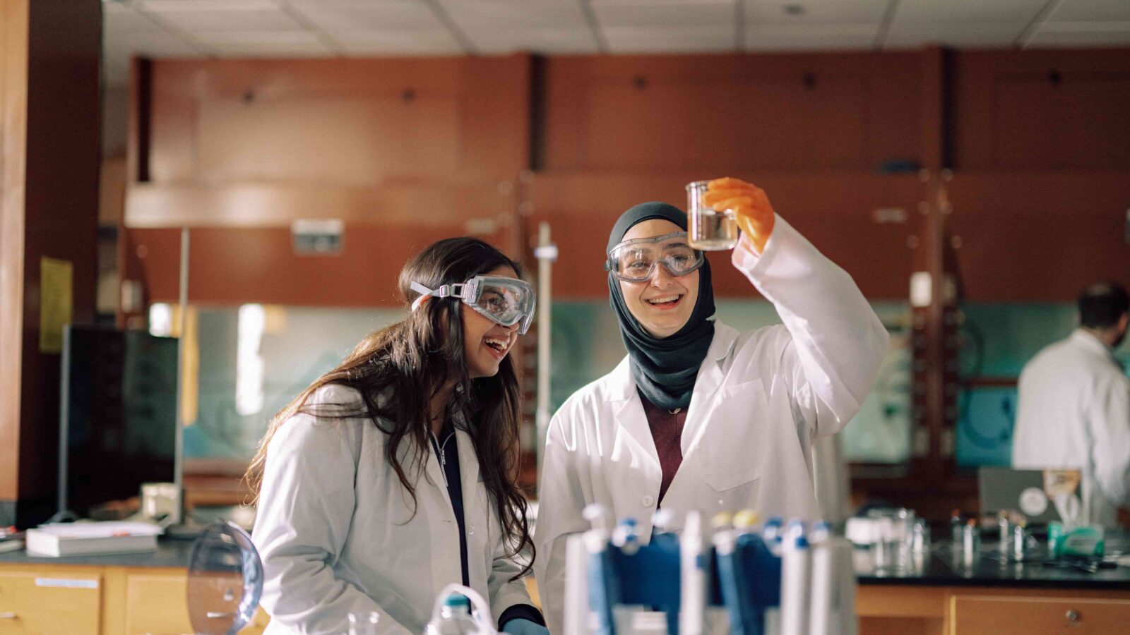 Two students in safety glasses and lab coats examining an experiment in a lab.