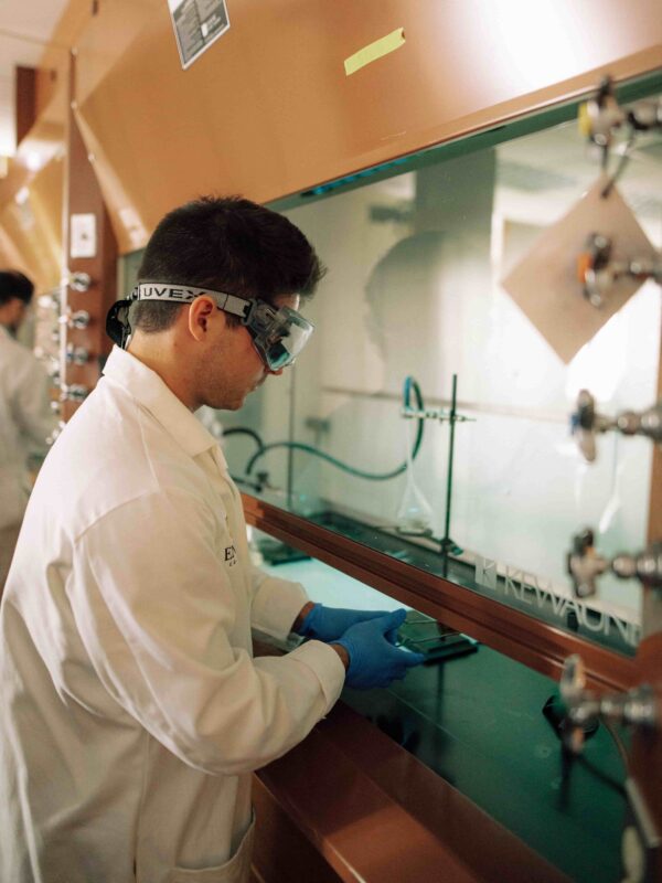 A student wearing safety glasses and a lab coat performing an experiment under a lab hood.