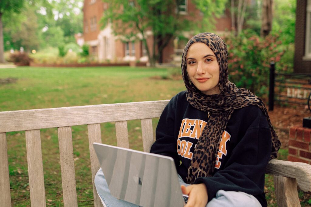 A student wearing a head scarf, sitting on an outdoor bench with a laptop in their lap.