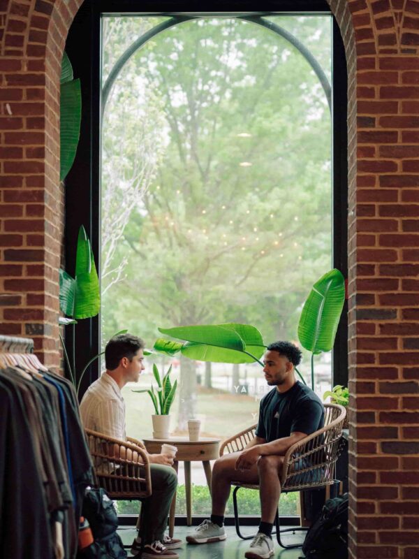 Two students chatting over coffee inside a building with brick columns.