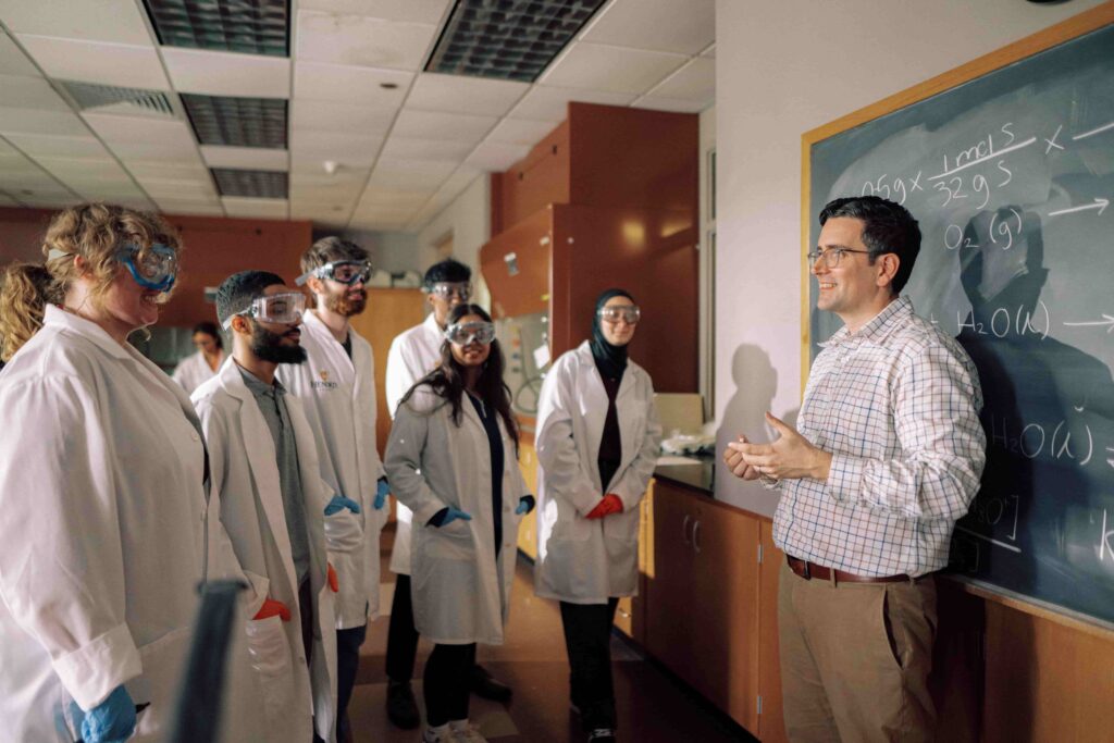 An instructor standing in front of a group of students wearing safety equipment in a science lab.