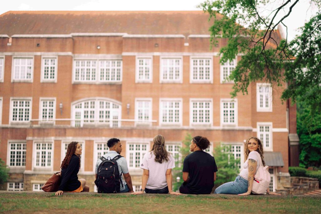 A group of five students sitting on a stone wall in front of a large brick building on the Hendrix campus.
