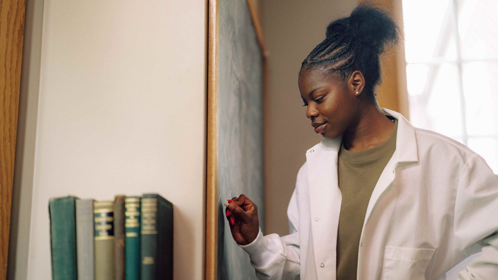 A student in a lab coat writing equations on a chalkboard.