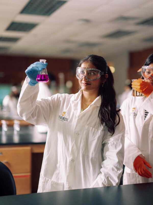 A student in safety goggles and lab coat examining the contents of a beaker in a science lab.