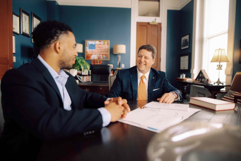 Two men in suits having a conversation at a table in an office with blue walls.