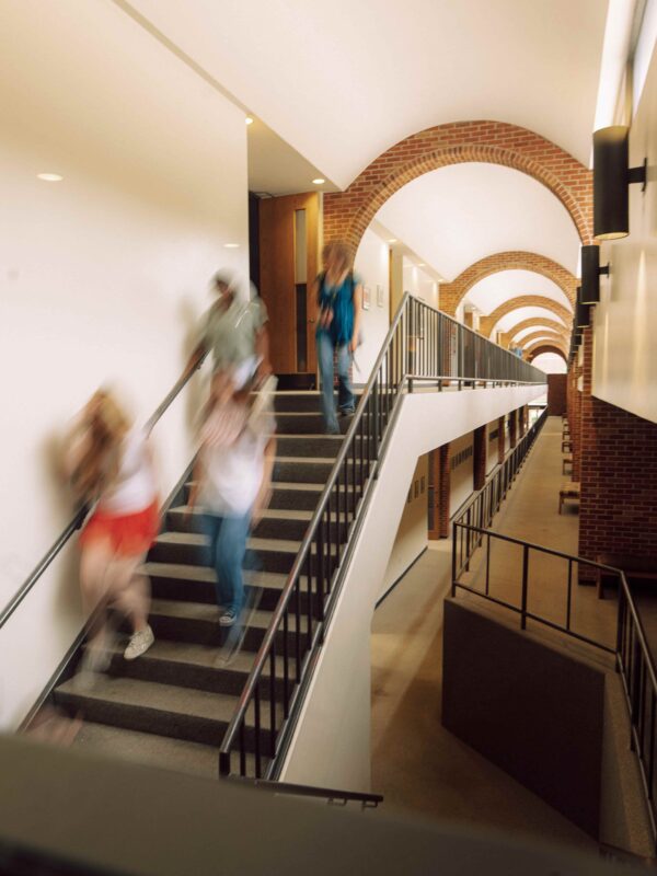 Students descending a staircase at the end of a long arched hallway.
