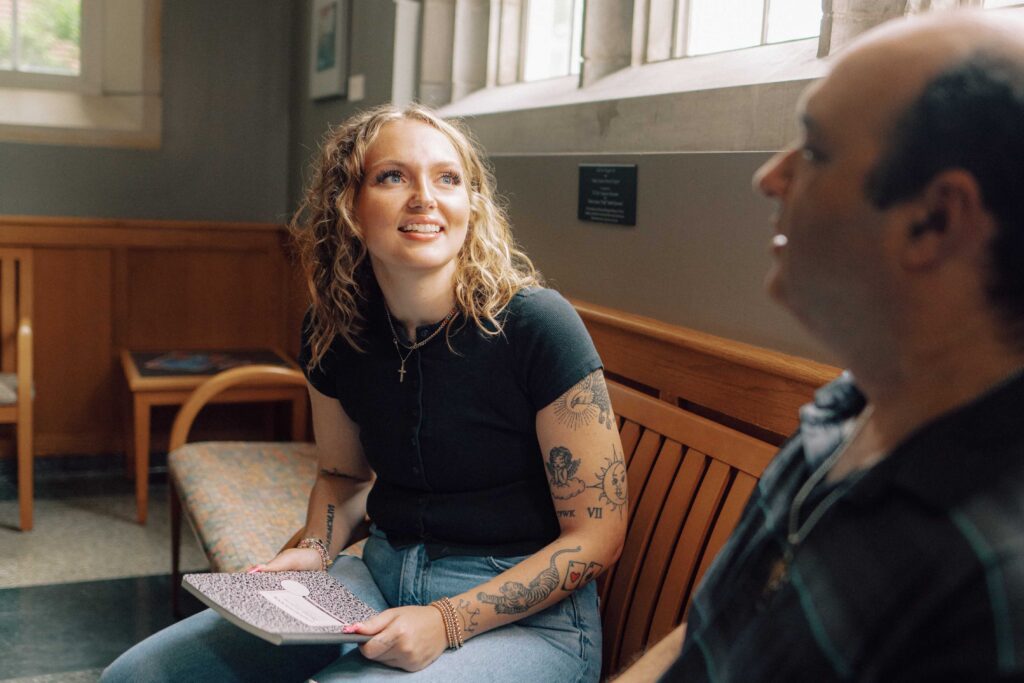 A student and a faculty member having a conversation on a wooden bench in a building on campus.