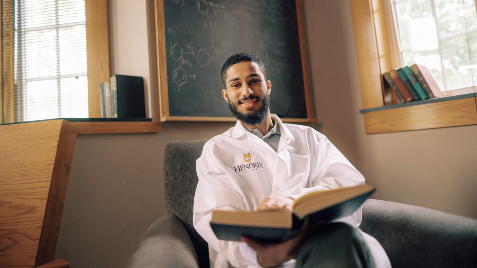 Smiling health science student in a Hendrix College lab coat holding a book in a classroom.
