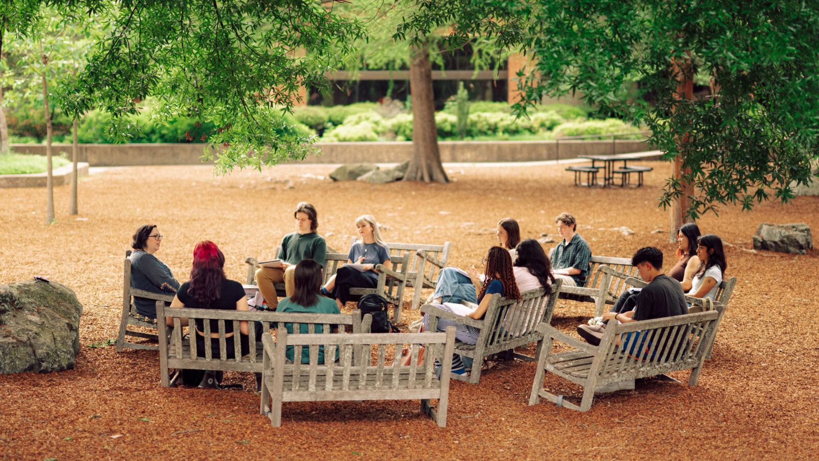 A Hendrix College professor leads an outdoor discussion with a class of students seated on benches under trees.