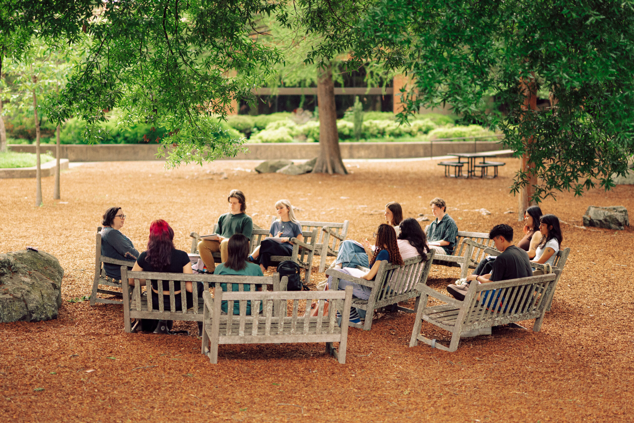 A Hendrix College professor leads an outdoor discussion with a class of students seated on benches under trees.