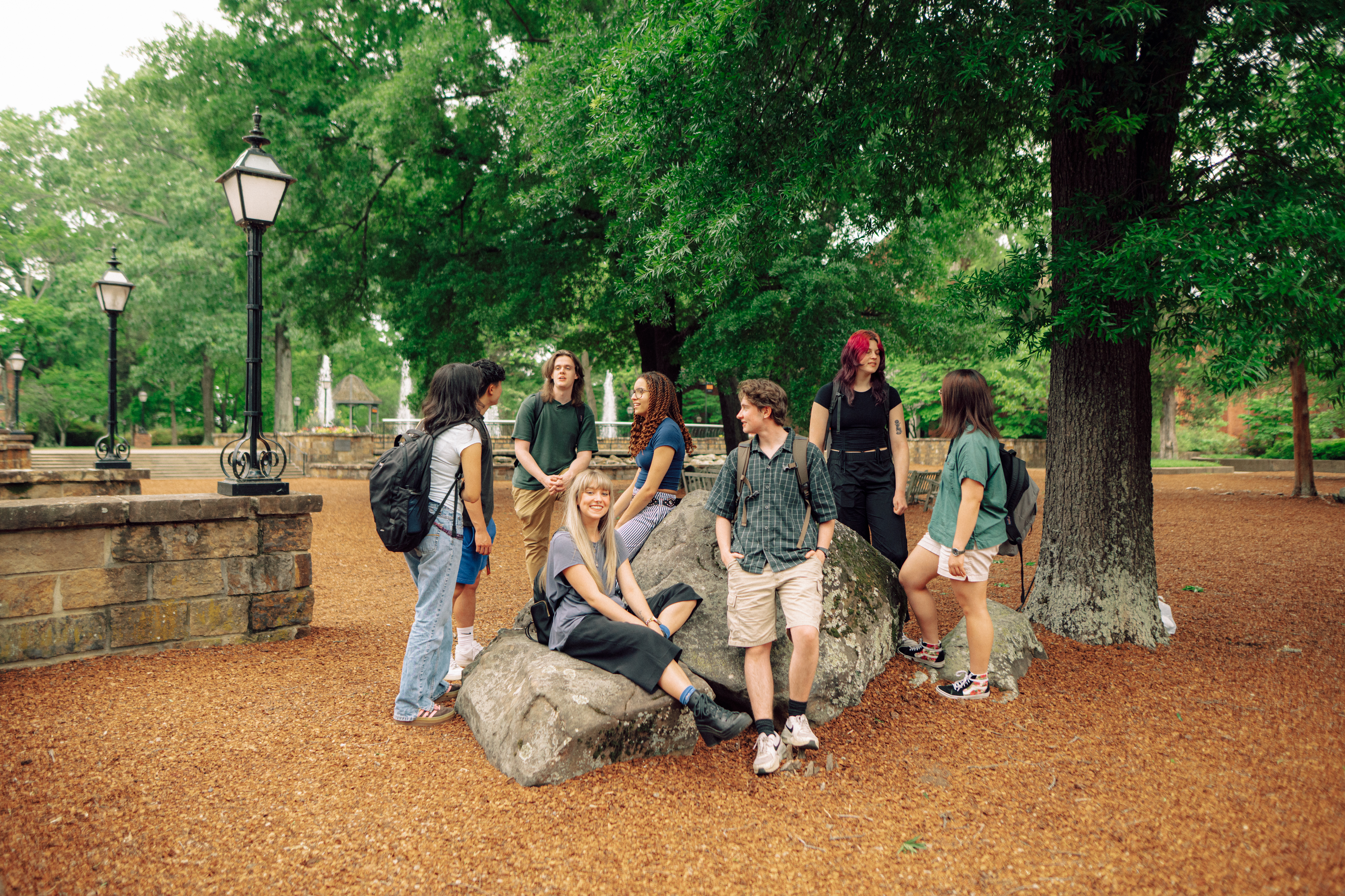 A group of Hendrix College students gather and talk outdoors seated on rocks on a wooded college campus plaza.