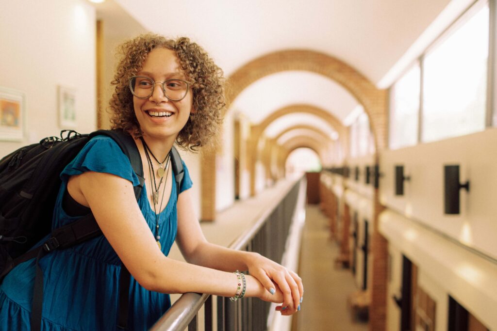 A smiling student leaning on a railing in a long arched hallway.