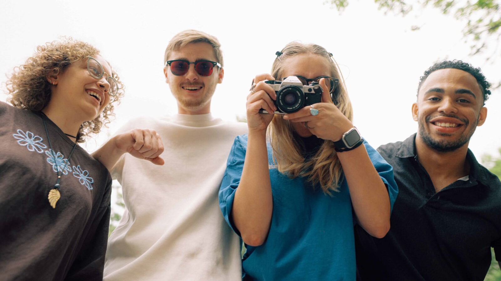 Four smiling Hendrix interdisciplinary students outdoors look down toward the camera as one points a film camera at the lens.