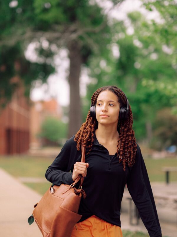 A student walking down the sidewalk in campus carrying a shoulder bag and wearing headphones.