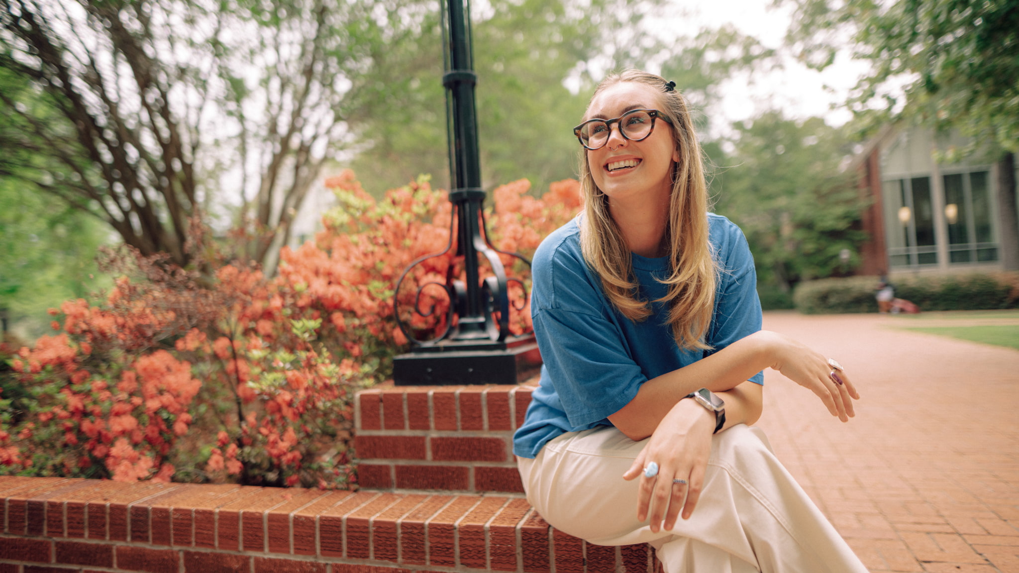 A smiling Hendrix College student sits on a brick ledge outside near orange flowers on a sunny campus pathway.