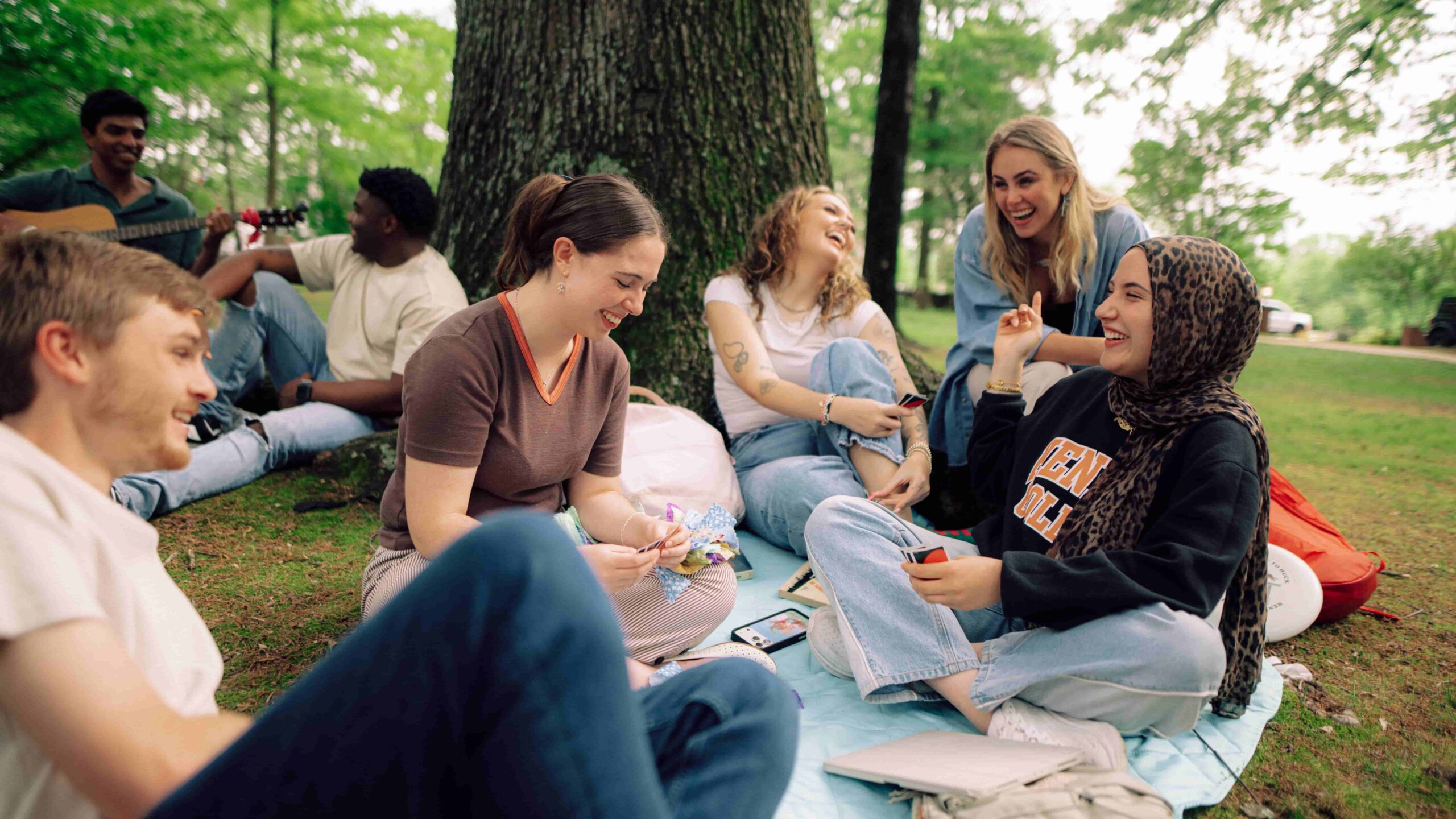 A group of Hendrix College students laugh, talk, and play card games on a blanket outside under trees on campus.
