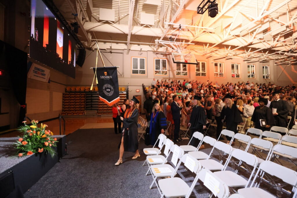 A graduate in a black cap and gown leads the commencement procession out of the crowded gymnasium, carrying a black and orange banner that reads "Hendrix College."