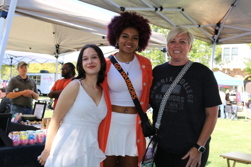 Three people smiling while standing under a tent on a sunny day outdoors.